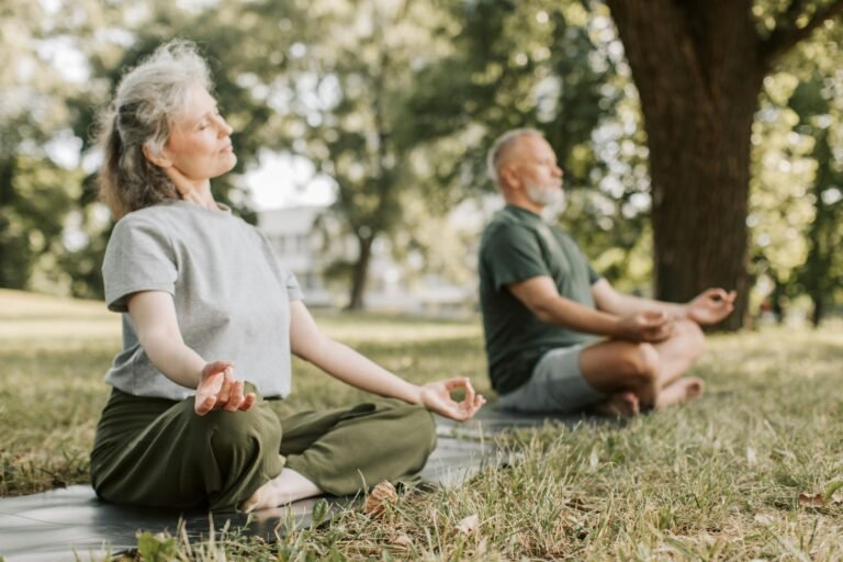 Elderly couple practicing meditation on yoga mats in a serene park setting.